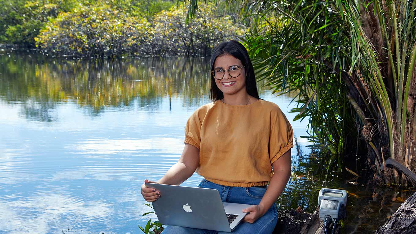 Mulher estuda em computador a margem de uma lagoa.