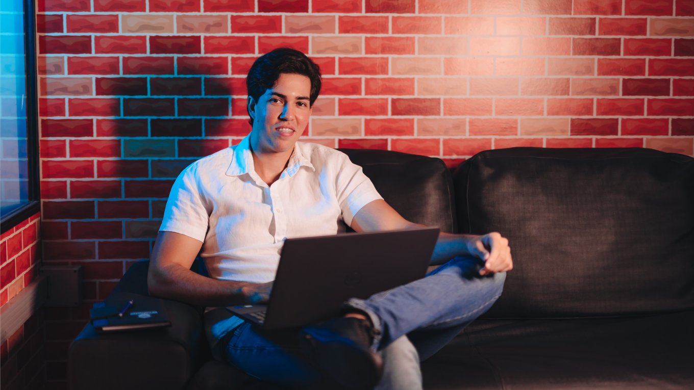 Jovem está sentando com notebook sobre as pernas; ele veste camisa branca e calça jeans