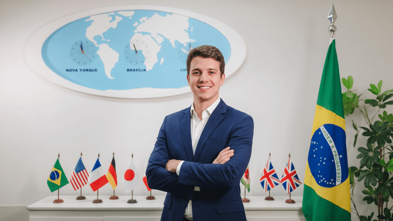 Um jovem rapaz está de blaser azul e se braços cruzados, em pé. Atrás dele há um mapa mundi e bandeiras de vários países. À sua direita, há uma bandeira do Brasil