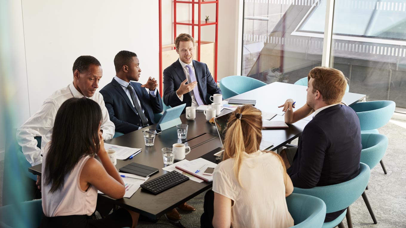 Seis profissionais debatem seriamente em torno de uma mesa de reunião.