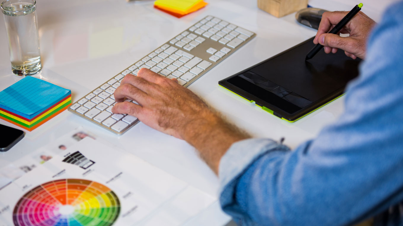 Mão em evidência mexe em teclado sobre mesa com tablet e post it.